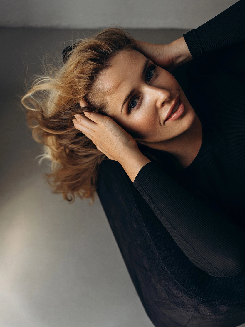 Woman with wavy blonde hair in black turtleneck posing with hands in hair against gray background - Brow lift in Newport Beach, CA