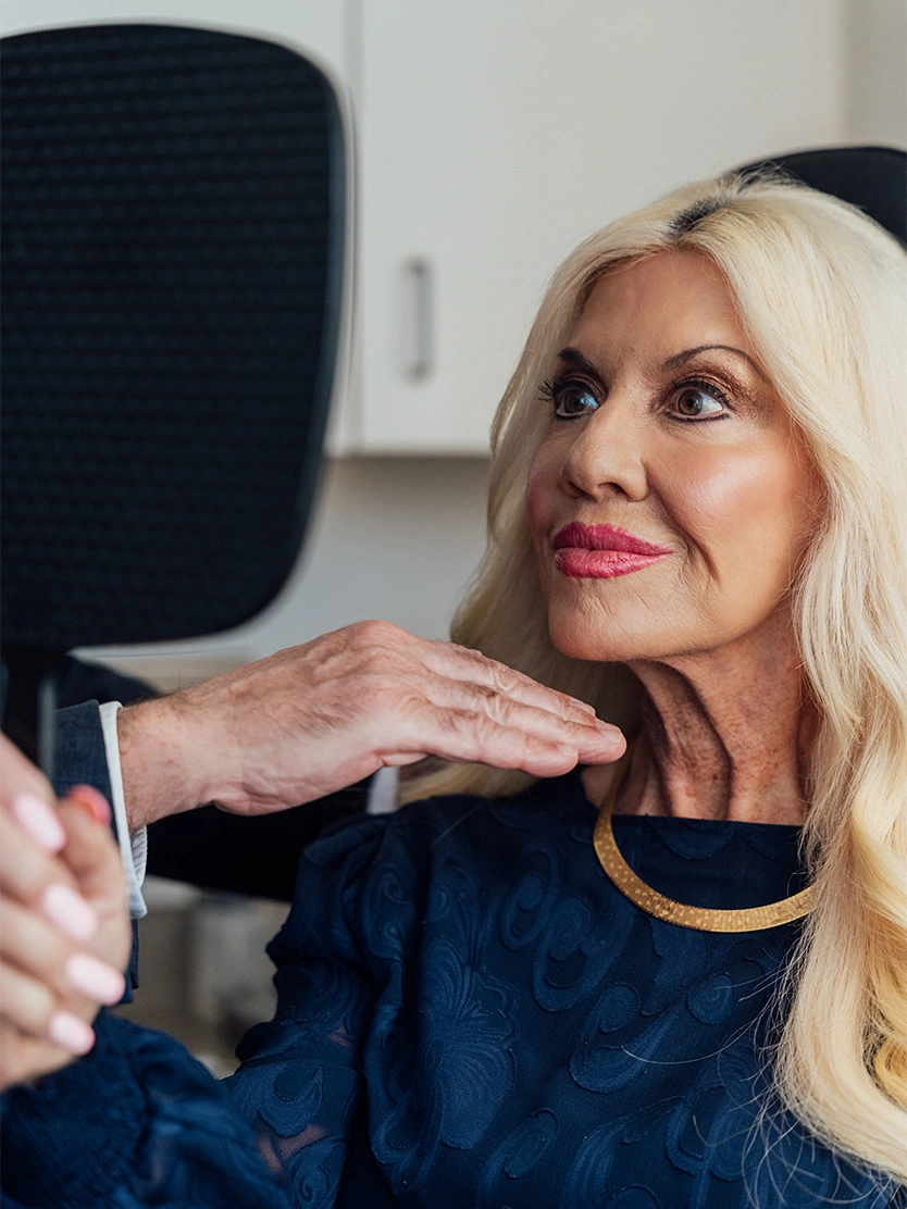 Mature blonde woman examining her face in a hand mirror while touching her chin in a modern office. - Deep plane neck lift in Newport Beach, CA