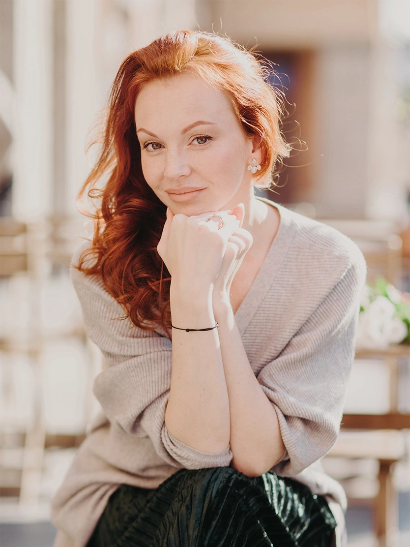 Red-haired woman in white blouse with hands clasped under chin, soft natural lighting portrait - Facelift after weight loss in Newport Beach, CA