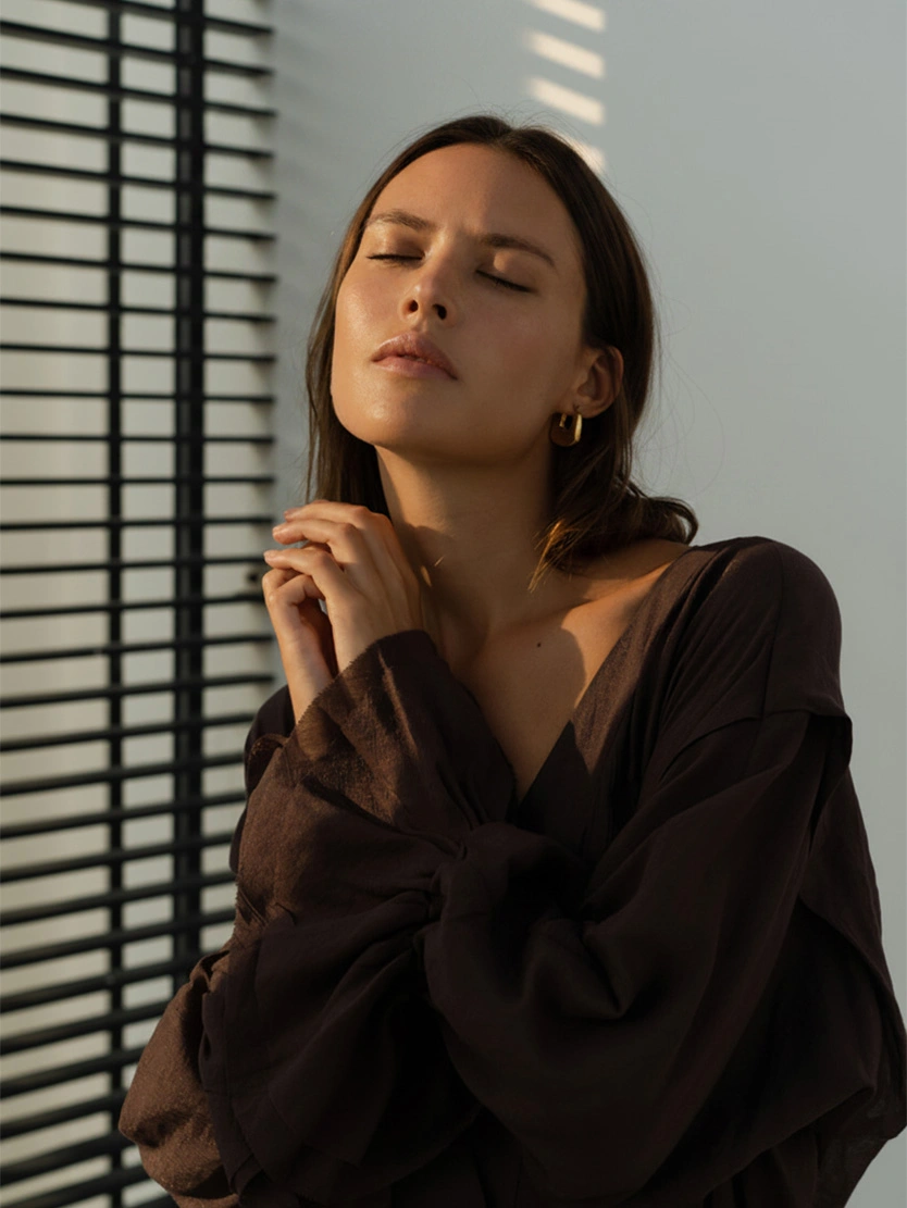 Woman in black blouse with eyes closed by window blinds creating shadow patterns in natural light - Liquid rhinoplasty in Newport Beach, CA
