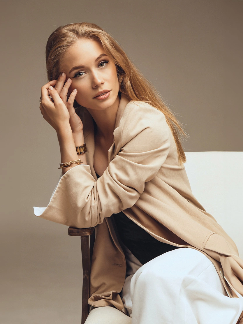 Woman in beige blouse and white pants sitting on chair in elegant studio portrait with neutral background - Sculptra in Newport Beach, CA