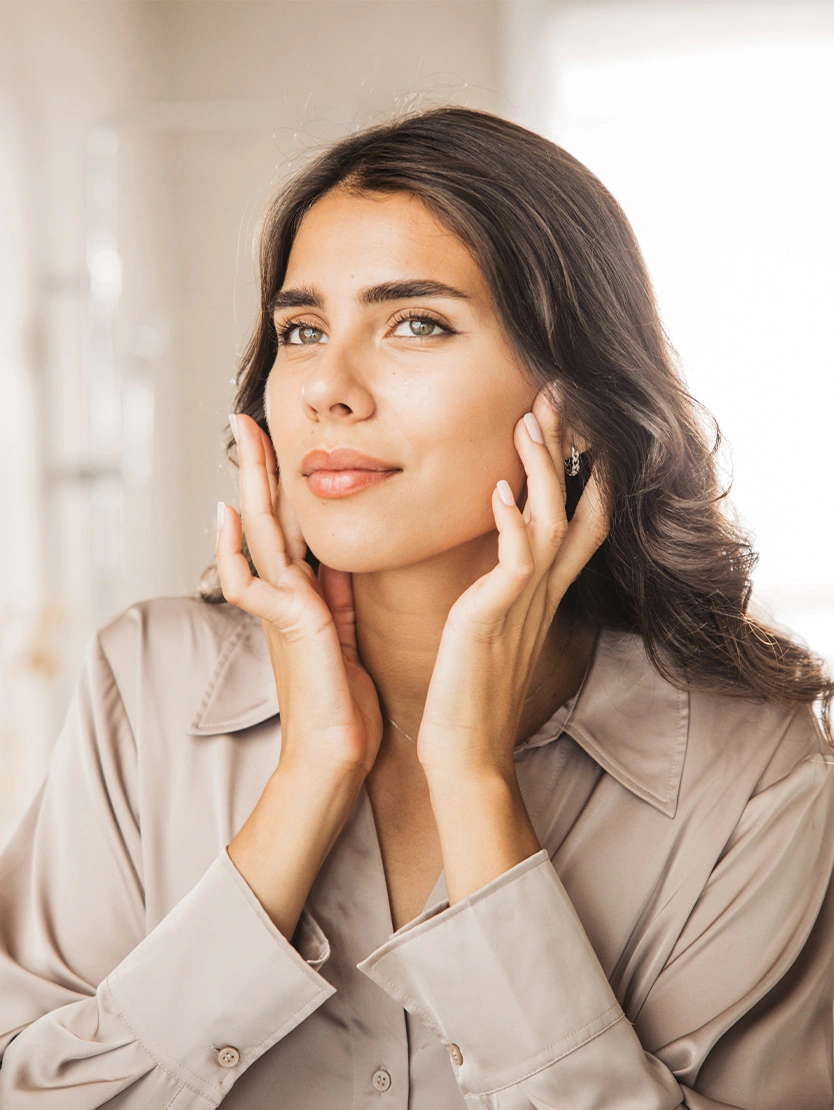 Woman with glowing skin touching face gently, demonstrating skincare routine and natural beauty in soft lighting - Skin boosters in Newport Beach, CA