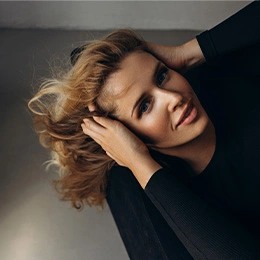 Woman with blonde wavy hair tilting head back with hands in black sleeves against gray background - Brow lift in Newport Beach, CA