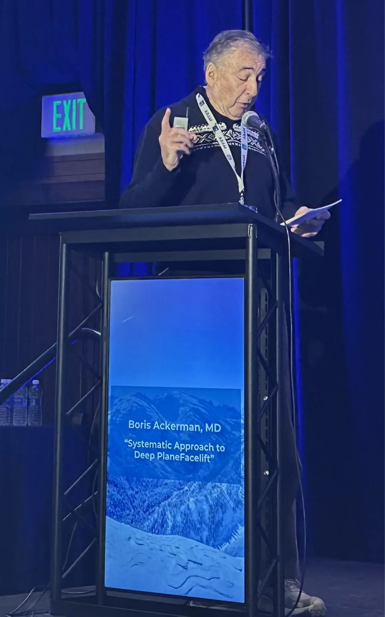 Dr Boris M Ackerman MD presenting at podium during conference with blue stage lighting and digital display screen