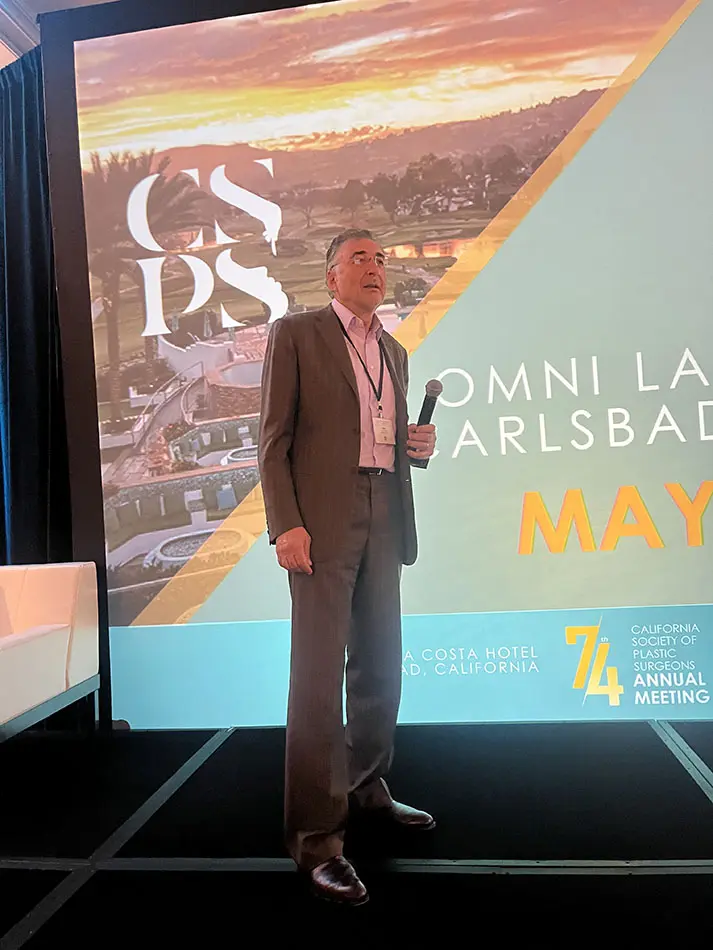 Dr Boris M Ackerman MD in suit standing on stage in front of presentation screen at MNI Laboratories Carlsbad event