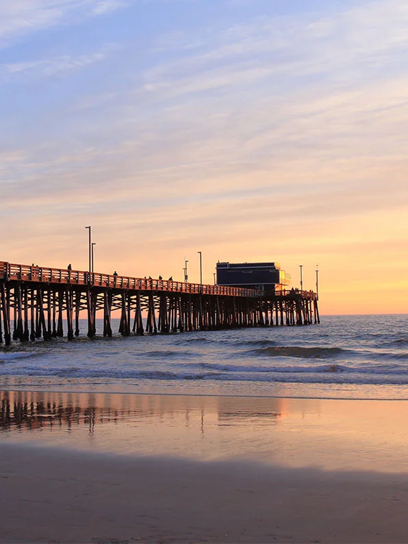 Sunset over Newport Beach Pier with wooden pilings reflected on wet sand and ocean waves