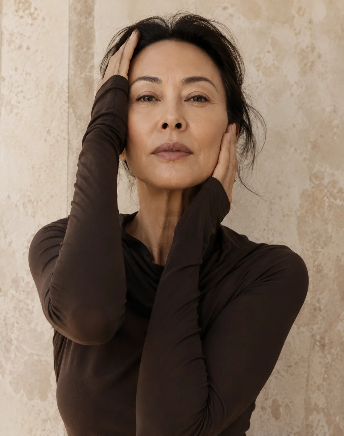 Woman in black turtleneck with hands in hair wearing white earrings against textured beige wall - Vertical facelift in Newport Beach, CA